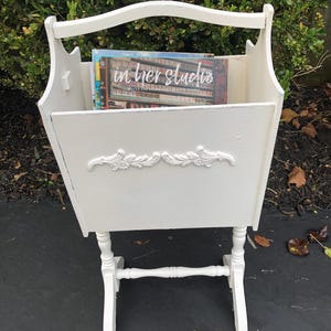 May include: White painted magazine rack with a handle and decorative floral accents. The rack is filled with magazines, and the text "in her studio" is visible on the cover of one magazine. The rack stands on four legs.
