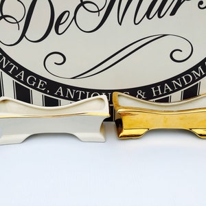 Op de afbeelding: Twee keramische potten, een wit met gouden rand en een gouden. Beide potten hebben een gebogen, langwerpige vorm met een lichte dip in het midden. De potten staan op een wit oppervlak.