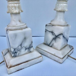 Puede incluir: Dos l&aacute;mparas de mesa de m&aacute;rmol blanco con vetas negras. Las l&aacute;mparas tienen una base cuadrada, un cuerpo en forma de columna y una parte superior decorativa. Las l&aacute;mparas est&aacute;n conectadas a una fuente de alimentaci&oacute;n con un cable marr&oacute;n. Las l&aacute;mparas est&aacute;n sobre una superficie blanca.