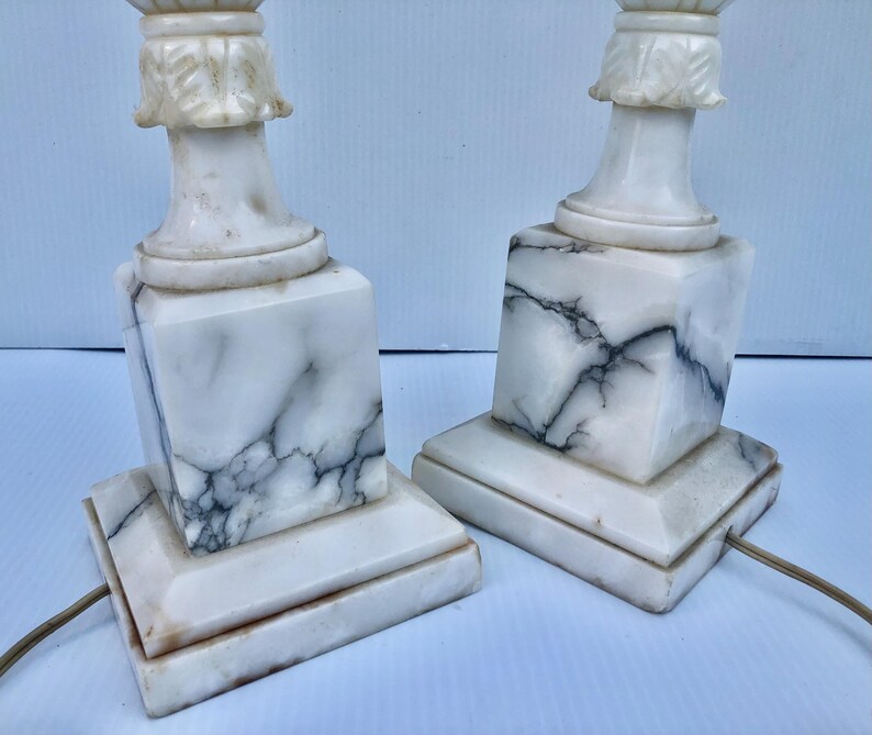 Puede incluir: Dos l&aacute;mparas de mesa de m&aacute;rmol blanco con vetas negras. Las l&aacute;mparas tienen una base cuadrada, un cuerpo en forma de columna y una parte superior decorativa. Las l&aacute;mparas est&aacute;n conectadas a una fuente de alimentaci&oacute;n con un cable marr&oacute;n. Las l&aacute;mparas est&aacute;n sobre una superficie blanca.