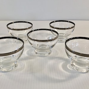 Pode incluir: Conjunto de cinco tigelas de sobremesa de vidro transparente com aro prateado. As tigelas têm uma base com pé.