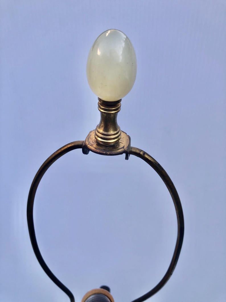 Puede incluir: Un remate de l&aacute;mpara vintage con una parte superior p&aacute;lida en forma de huevo. El remate est&aacute; montado sobre una base de lat&oacute;n con un marco de metal curvado. El dise&ntilde;o es simple y elegante, adecuado para una l&aacute;mpara cl&aacute;sica o antigua.