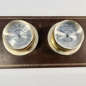 Puede incluir: Una estación meteorológica vintage con termómetro e higrómetro. Los instrumentos tienen esferas plateadas con marcas negras y carcasas de metal dorado. Están colocados sobre una base marrón similar al cuero con un borde dorado decorativo. La palabra "SPRINGFIELD" es visible.