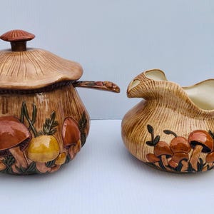 May include: A ceramic mushroom-themed serving set. The set includes a lidded bowl and a creamer jug. The bowl has a mushroom-shaped lid and is decorated with brown, yellow, and orange mushrooms. The creamer has a handle and is also decorated with mushrooms.