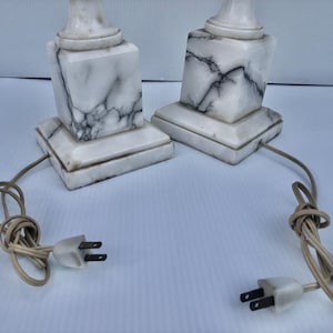 Puede incluir: Dos l&aacute;mparas de m&aacute;rmol a juego con un patr&oacute;n veteado blanco y gris. Cada l&aacute;mpara tiene una base cuadrada y una parte superior cil&iacute;ndrica. Las l&aacute;mparas est&aacute;n conectadas a cables el&eacute;ctricos con enchufes de dos clavijas.