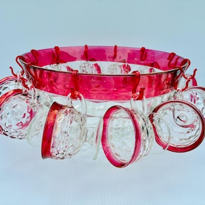 Puede incluir: Un cuenco de cristal transparente con un borde ancho de color rojo intenso y una base texturizada con patrón de diamantes. Varias tazas pequeñas de cristal a juego con bordes rojos cuelgan del borde del cuenco mediante ganchos metálicos.