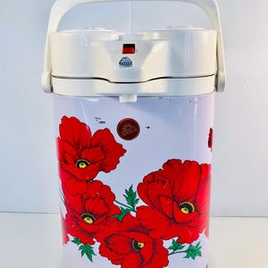 Puede incluir: Una caldera de agua floral blanca y roja con un asa blanca. La caldera de agua tiene un interruptor rojo y negro y un logotipo "Superior" rojo y dorado. El diseño floral presenta grandes amapolas rojas con hojas verdes.