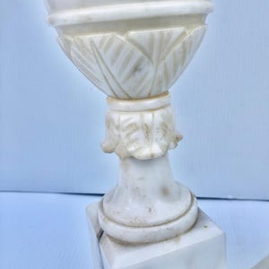 Puede incluir: Un par de urnas decorativas de m&aacute;rmol blanco. Las urnas tienen un dise&ntilde;o detallado con una parte superior acanalada y una base con tallas en forma de hoja. Las urnas se asientan sobre una base cuadrada. El m&aacute;rmol tiene vetas grises.