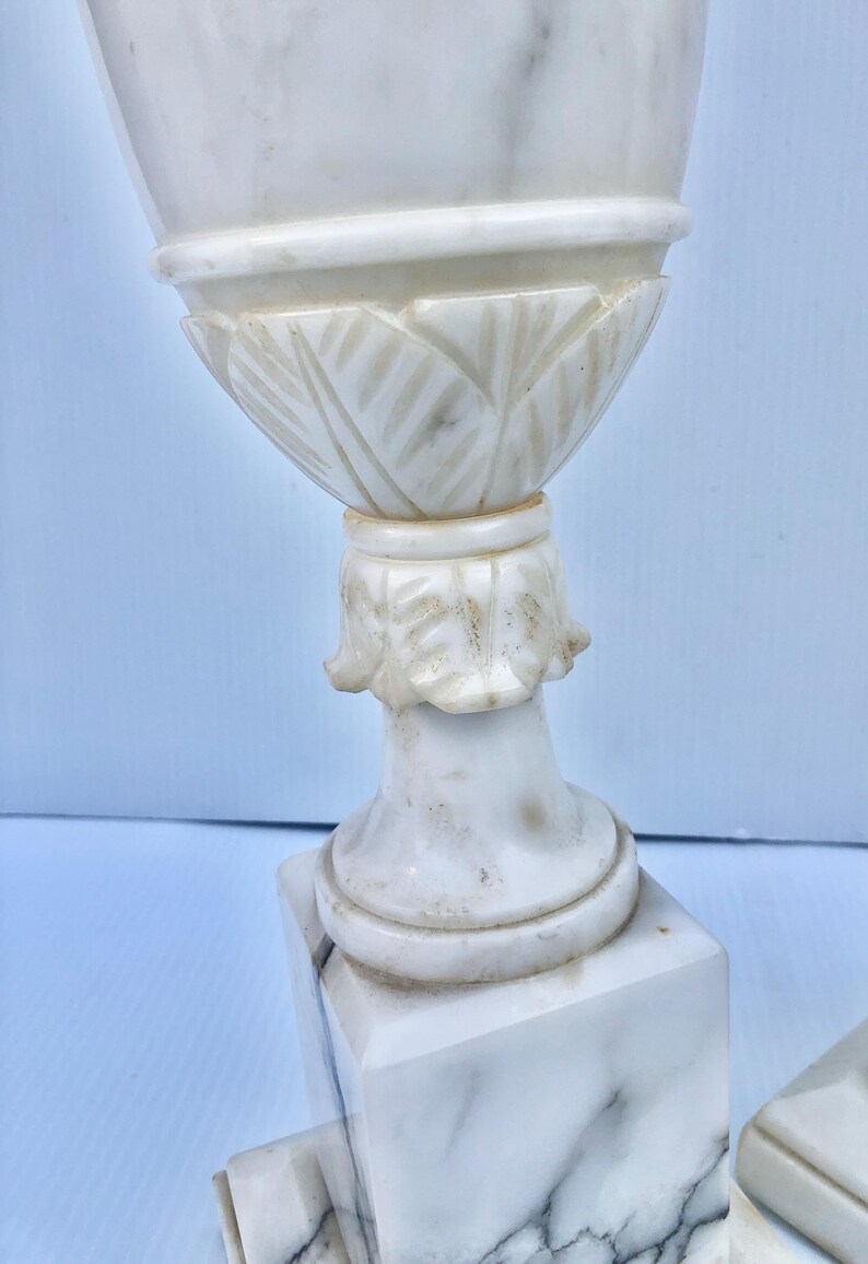 Puede incluir: Un par de urnas decorativas de m&aacute;rmol blanco. Las urnas tienen un dise&ntilde;o detallado con una parte superior acanalada y una base con tallas en forma de hoja. Las urnas se asientan sobre una base cuadrada. El m&aacute;rmol tiene vetas grises.