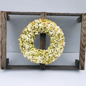 May include: A decorative wreath made of cream and yellow flowers, centered on a rectangular frame. The frame is constructed of weathered wood and wire mesh, creating a rustic aesthetic. The wreath is a circular design, perfect for seasonal home decor.
