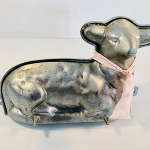 May include: A vintage silver-coloured metal lamb cake mould, with a light pink ribbon tied around its neck. The lamb has detailed features and is designed for baking a lamb-shaped cake. Ideal for Easter or spring celebrations.