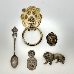 May include: An assortment of vintage metal lion-themed decorative items. Includes a door knocker, a spoon with a crest, a lion head, a lion figurine, and a pendant. The items are in various shades of gold, silver, and bronze.