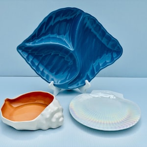 May include: A collection of three ceramic dishes: a large, deep blue, shell-shaped divided tray, a white shell-shaped bowl with a vibrant orange interior, and a flat, iridescent white shell-shaped plate with a textured, radiating ribbed surface.