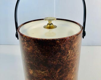 May include: A vintage ice bucket with a brown and gold mottled finish. It has a white interior, a gold-toned knob on the lid, and a black handle. The bucket is cylindrical in shape.