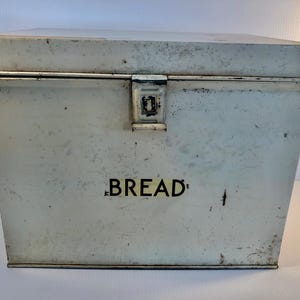 Op de afbeelding: Witte metalen broodtrommel met een scharnierend deksel en een metalen sluiting. De trommel heeft een versleten, verweerde afwerking en het woord "BREAD" is op de voorkant gedrukt.
