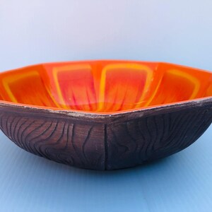 Könnte beinhalten: Eine Keramikschale mit einzigartigem Design. Der Innenraum ist leuchtend orange mit gelben Akzenten, während die Außenseite eine dunkelbraune Holzmaserung aufweist. Die Schale hat eine geometrische Form, ideal zum Servieren oder Dekorieren.