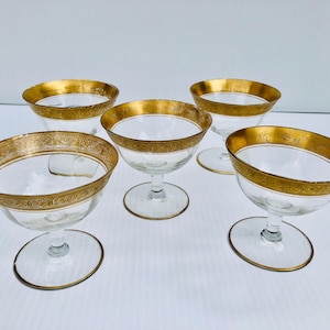 May include: Five clear champagne coupe glasses with gold rims and bases. The gold rims have an ornate pattern. The glasses are on a white surface, highlighting their elegant design. Each glass has a gold rim.