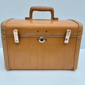 May include: A rectangular tan hard-shell train case with a smooth finish, featuring a sturdy tan top handle, two silver-toned metal latches on the front, and a central circular keyhole mechanism. The case has rounded edges and a structured design.