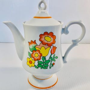 Puede incluir: Tetera de cerámica blanca con un diseño floral en naranja, amarillo y verde. La tetera tiene un pico curvo, un asa de bucle y una tapa con un adorno decorativo. Ribete naranja en la base y la tapa.