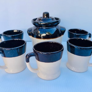 Puede incluir: Un juego de seis tazas de cerámica marrón y blanca con una jarra de cerámica marrón y blanca a juego. Las tazas tienen una banda marrón alrededor de la parte superior y una banda blanca alrededor de la parte inferior. La jarra tiene una tapa marrón.