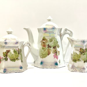May include: A white porcelain tea set with a floral design. The set includes a teapot, creamer, and sugar bowl. The floral design features green leaves and pink flowers.