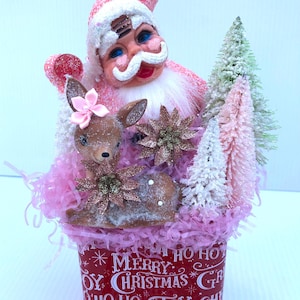 May include: Pink and white Santa Claus figurine with a pink reindeer and pink and white Christmas trees in a red tin with the text "Merry Christmas" and "Ho Ho Ho" printed on it.