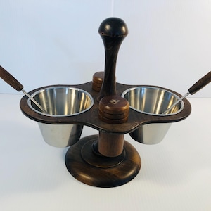 Könnte beinhalten: Ein Vintage-Gewürzständer mit einem dunklen Holzsockel und Griff. Er verfügt über zwei Edelstahlschalen, zwei kleine Holzbehälter und zwei Löffel. Der Ständer ist für Salz, Pfeffer und andere Gewürze konzipiert.