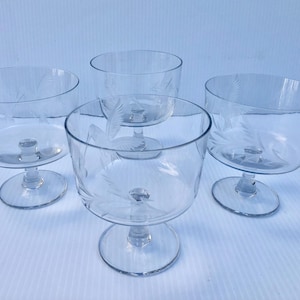 May include: Four clear glass dessert bowls with etched leaf designs. Each bowl has a rounded shape, a stem, and a circular base. The bowls are arranged on a light blue surface.