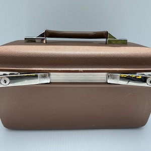 May include: A rectangular brown hard-shell travel case with a textured exterior finish. The case features a dark brown top handle, a silver-toned metal trim along the center seam, and two chrome-plated locking latches on the front side.