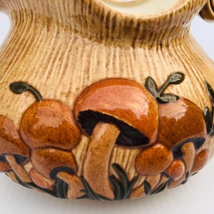 May include: A ceramic jug with a textured, light brown exterior. The jug features a relief design of brown and orange mushrooms with green leaves. The handle and spout are also visible.