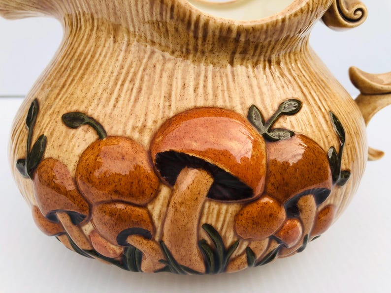 May include: A ceramic jug with a textured, light brown exterior. The jug features a relief design of brown and orange mushrooms with green leaves. The handle and spout are also visible.