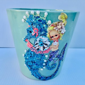 May include: A turquoise ceramic planter with a mermaid and seahorse design. The mermaid is holding a pearl and the seahorse is decorated with blue glitter and pearls.