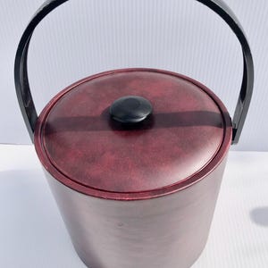 May include: A burgundy ice bucket with a black handle and lid. The cylindrical bucket has a matching burgundy lid with a black knob. The black handle arches over the top of the bucket.