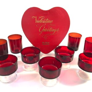 Peut inclure: Ensemble de dix verres en verre rouge, dont six petits verres à pied et quatre verres plus hauts. Les verres sont disposés sur une surface blanche avec une boîte en forme de cœur rouge qui dit "Valentine Greetings" en lettres dorées.