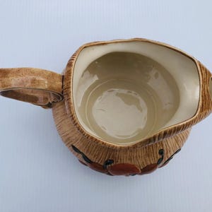 May include: A brown ceramic jug with a handle and a wide opening. The jug has a textured exterior with a design of red and black accents. The interior is a light beige colour.