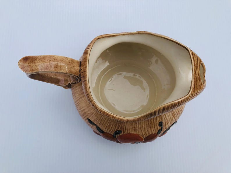 May include: A brown ceramic jug with a handle and a wide opening. The jug has a textured exterior with a design of red and black accents. The interior is a light beige colour.