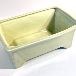 May include: A pale green ceramic rectangular planter with a slightly raised rim and three small feet on the bottom.