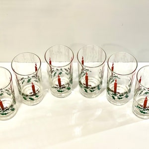 Peut inclure: Six verres transparents orn&eacute;s d'un motif festif de bougie et de houx de No&euml;l. Chaque verre pr&eacute;sente une bougie rouge avec une flamme et une couronne de houx verte. Les verres sont dispos&eacute;s sur une surface blanche.