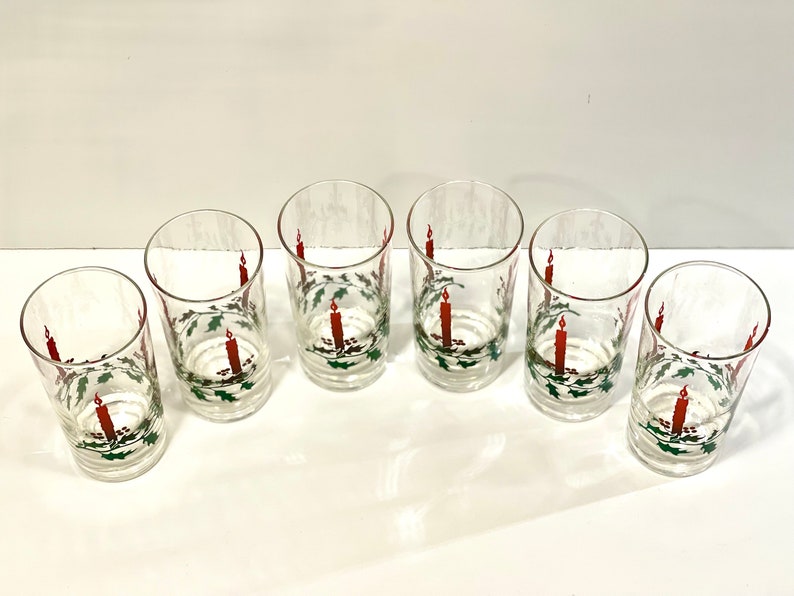 Peut inclure: Six verres transparents orn&eacute;s d'un motif festif de bougie et de houx de No&euml;l. Chaque verre pr&eacute;sente une bougie rouge avec une flamme et une couronne de houx verte. Les verres sont dispos&eacute;s sur une surface blanche.
