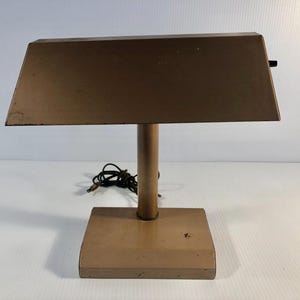 Puede incluir: Lámpara de escritorio vintage de metal color bronce. Cuenta con una pantalla rectangular y una base cilíndrica. La lámpara está diseñada para colocarse sobre un escritorio o mesa y proporcionar iluminación enfocada.