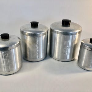 Puede incluir: Juego de cuatro botes de cocina vintage de aluminio con pomos negros. Los botes están etiquetados con las palabras: "Harina", "Azúcar", "Café" y "Té". Los botes son cilíndricos y tienen un acabado de metal cepillado.