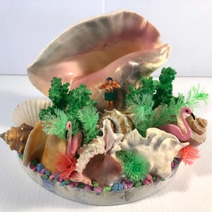 Pode incluir: Arranjo decorativo de conchas com uma grande concha como pano de fundo. O arranjo inclui várias conchas, plantas de plástico verde, uma dançarina de hula em miniatura e figuras de flamingos cor-de-rosa. A base é uma tigela de cerâmica com seixos coloridos.