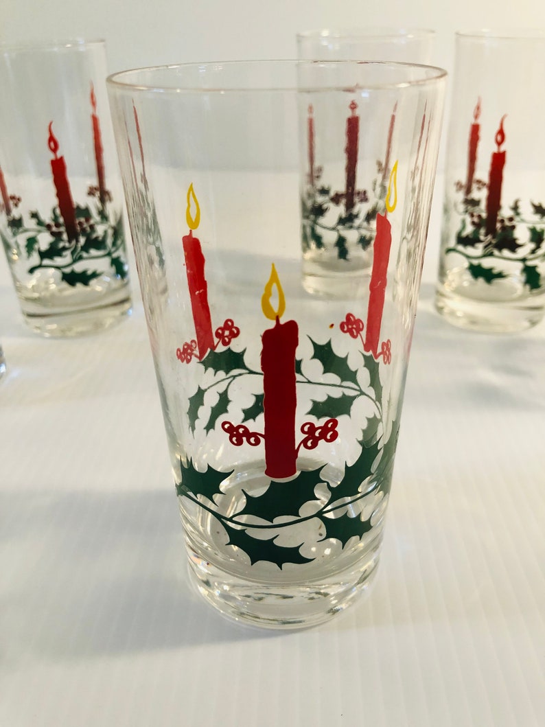 Peut inclure: Verres &agrave; boire transparents avec un motif de No&euml;l festif. Chaque verre pr&eacute;sente une bougie rouge avec une flamme jaune, entour&eacute;e de feuilles de houx vertes et de baies rouges. Les verres sont cylindriques et semblent &ecirc;tre en verre.