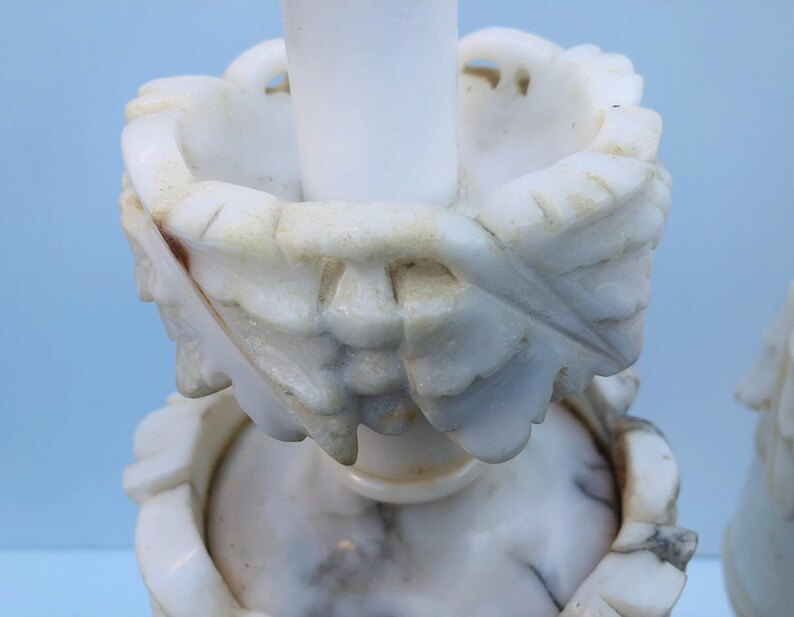Puede incluir: Un par de candelabros de m&aacute;rmol blanco. Los candelabros presentan intrincadas tallas de hojas y flores. El m&aacute;rmol tiene un aspecto veteado natural con sutiles variaciones de color, lo que se suma a su est&eacute;tica antigua.