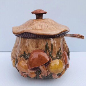 May include: A ceramic mushroom-shaped container with a lid and a handle. The container is primarily brown with detailed mushroom and leaf designs in shades of brown, orange, and yellow. The lid is shaped like a mushroom cap.
