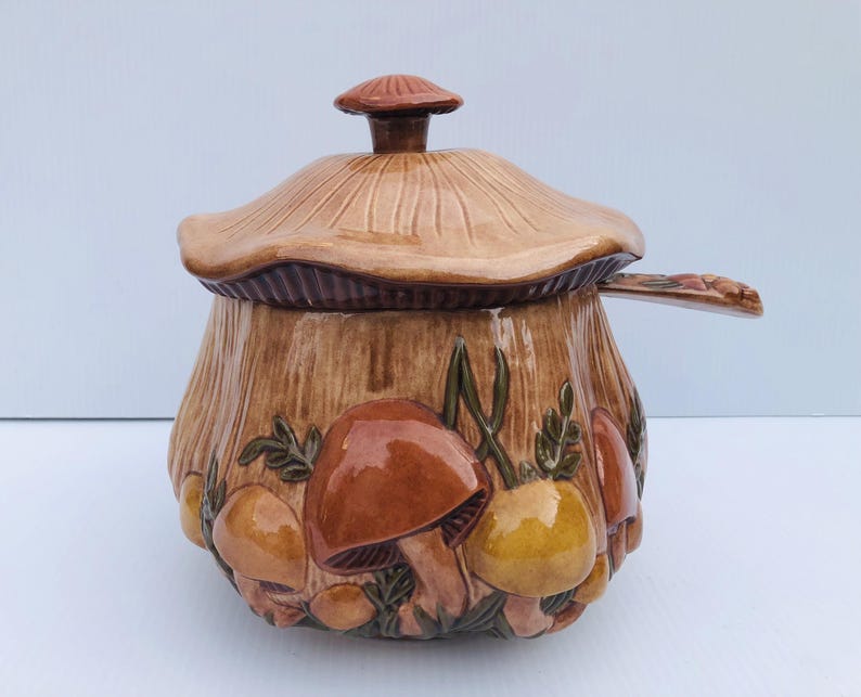 May include: A ceramic mushroom-shaped container with a lid and a handle. The container is primarily brown with detailed mushroom and leaf designs in shades of brown, orange, and yellow. The lid is shaped like a mushroom cap.