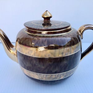 May include: A brown teapot with gold trim and a lid. The teapot has a round body and a spout on one side. The handle is on the opposite side of the spout.