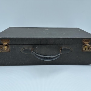 May include: A rectangular black suitcase with a textured exterior surface, a central black handle, and two brass-coloured metal latches with integrated keyholes positioned on the left and right sides of the front panel.