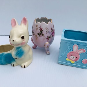 Könnte beinhalten: Drei Keramik-Blumentöpfe, einer in Form eines Eies mit Blumenmustern, einer in Form eines Hasen mit blauen Akzenten und ein quadratischer Topf mit blau-weißem Muster und einer rosa Hasenillustration.