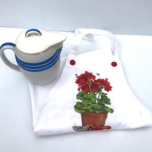 May include: A white apron with red buttons and a floral design featuring a red geranium in a pot, a bee, and garden shears. A ceramic pitcher with a lid and blue stripes is also in the image.